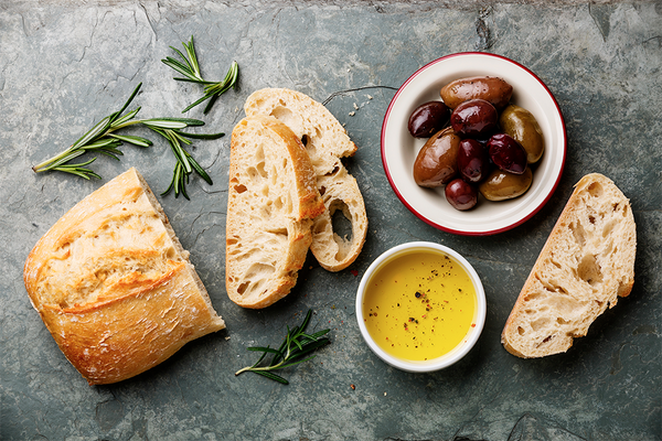 Pain coupé en tranche avec une branche de romarin, une coupelle d'olives noires et une coupelle d'huile d'olive